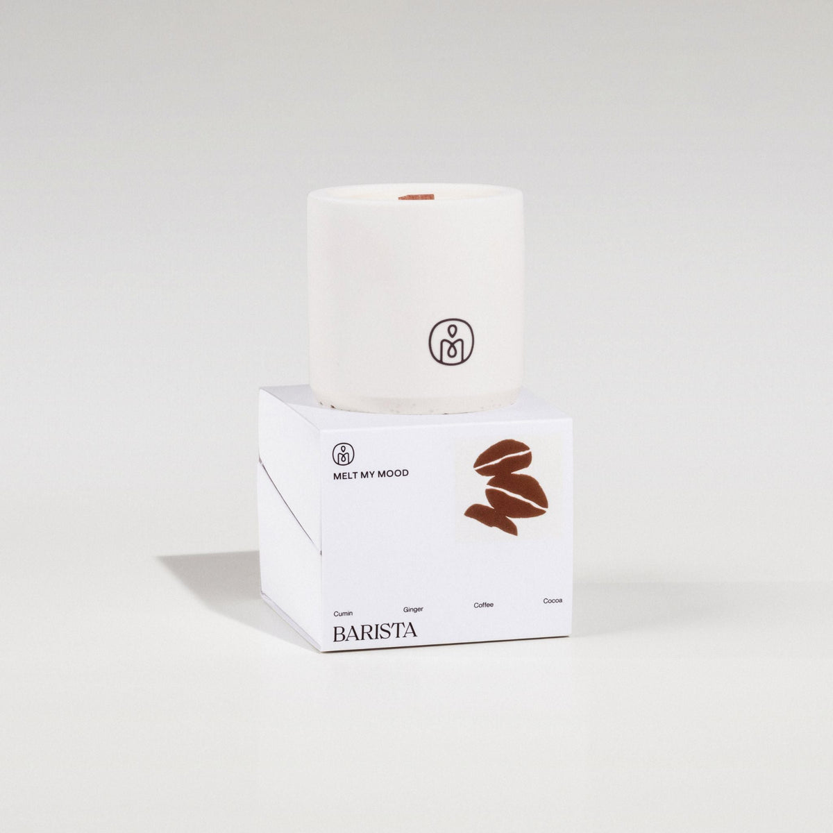 Egy henger alakú, törtfehér szójaviaszgyertya, benne fa kanóccal, egy fehér, kocka alakú dobozon áll. A dobozon a „BARISTA” (a gyertya neve) és egy barna kávészem ikon látható; a felvétel tiszta fehér stúdióháttér előtt, lágy árnyékkal készült.