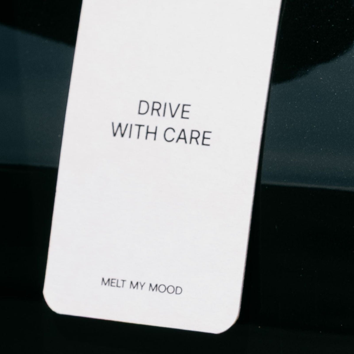 Lekerekített sarkú, fehér, téglalap alakú autóillatosító, amely fekete zsinóron lóg. Az autóillatosítón fekete felirat olvasható: „DRIVE WITH CARE”, alul pedig a „MELT MY MOOD” szöveg szerepel.