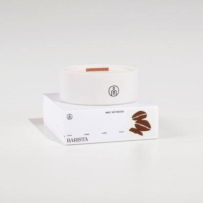 Egy kerek, törtfehér szójaviaszgyertya, benne fa kanóccal, egy fehér, téglalap alakú dobozon áll. A dobozon a „BARISTA” (a gyertya neve) és egy barna kávészem ikon látható; a felvétel tiszta fehér stúdióháttér előtt, lágy árnyékkal készült.