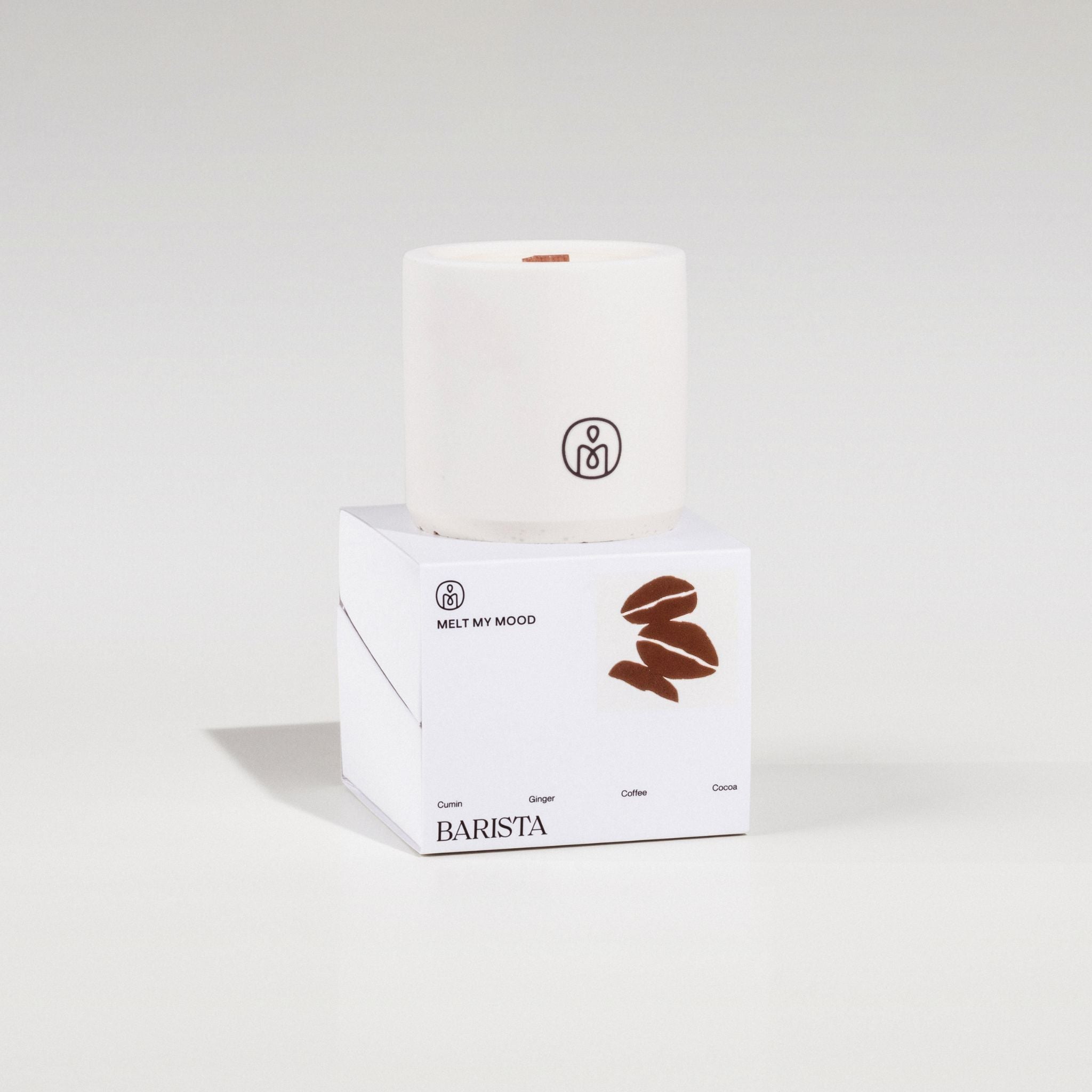 Egy henger alakú, törtfehér szójaviaszgyertya, benne fa kanóccal, egy fehér, kocka alakú dobozon áll. A dobozon a „BARISTA” (a gyertya neve) és egy barna kávészem ikon látható; a felvétel tiszta fehér stúdióháttér előtt, lágy árnyékkal készült.