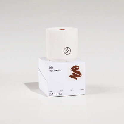 Egy henger alakú, törtfehér szójaviaszgyertya, benne fa kanóccal, egy fehér, kocka alakú dobozon áll. A dobozon a „BARISTA” (a gyertya neve) és egy barna kávészem ikon látható; a felvétel tiszta fehér stúdióháttér előtt, lágy árnyékkal készült.
