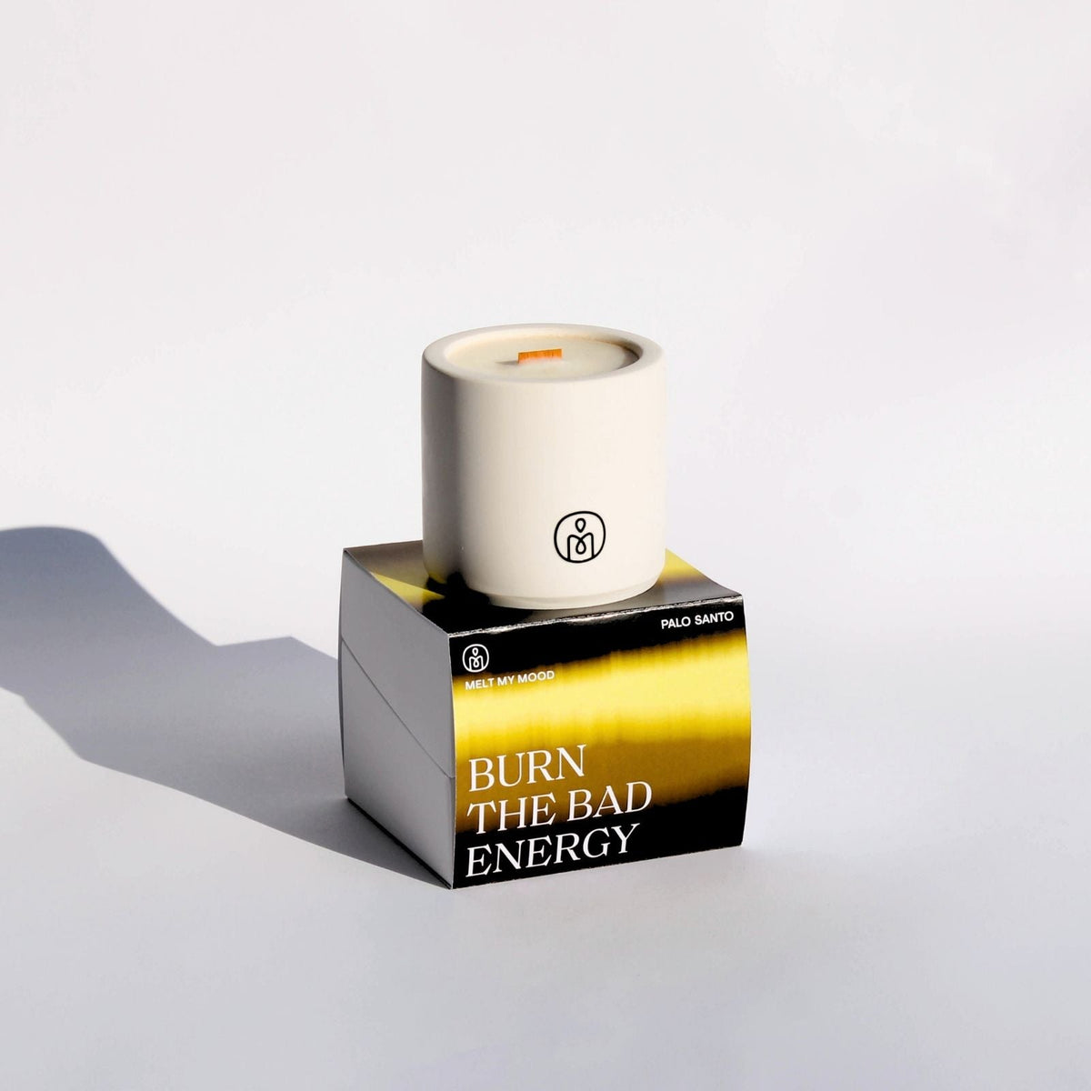 Egy henger, törtfehér gyertya, benne fa kanóccal, egy fekete, arany és zöld színű, kocka alakú dobozon áll. A dobozon a „BURN THE BAD ENERGY” (a gyertya neve) és a „PAOLO SANTO” (a gyertya illata) felirat látható; a felvétel tiszta fehér stúdióháttér előtt, lágy árnyékkal készült.