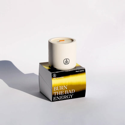 Egy henger, törtfehér gyertya, benne fa kanóccal, egy fekete, arany és zöld színű, kocka alakú dobozon áll. A dobozon a „BURN THE BAD ENERGY” (a gyertya neve) és a „PAOLO SANTO” (a gyertya illata) felirat látható; a felvétel tiszta fehér stúdióháttér előtt, lágy árnyékkal készült.