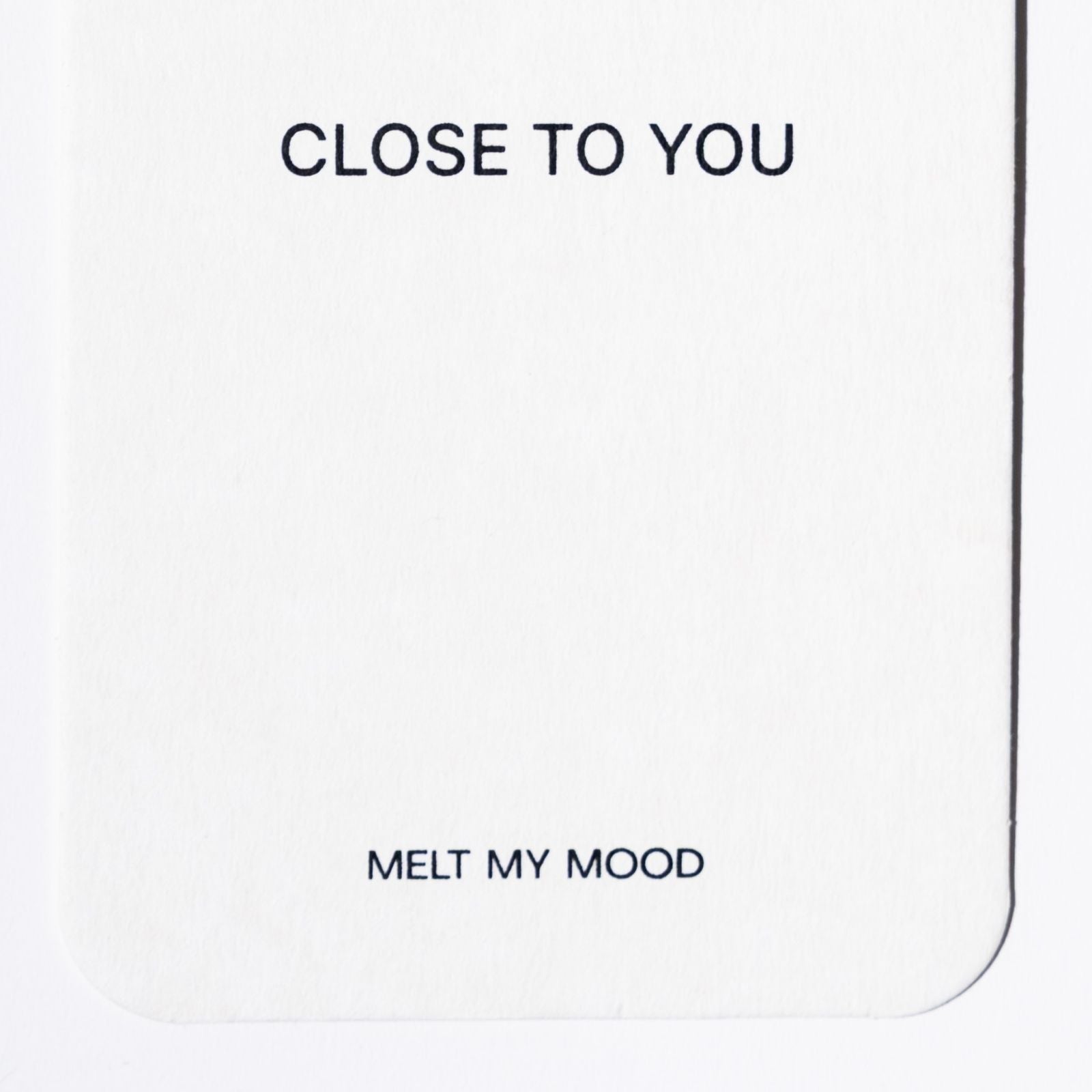 Közelkép egy lekerekített sarkú, fehér, téglalap alakú gardróbillatosítóról, amelynek felső részén a „CLOSE TO YOU”, alsó részén pedig a „MELT MY MOOD” felirat látható fekete betűkkel.