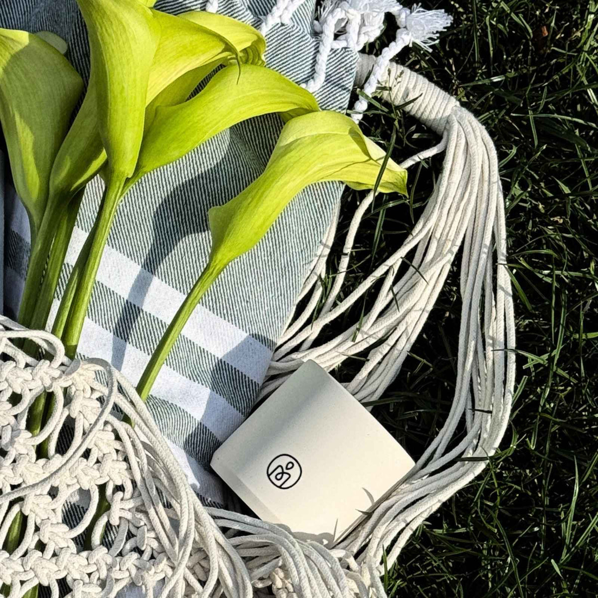 Egy fekete Melt My Mood logóval ellátott fehér gyertya fekszik egy fehér makramé táskában, fekete-fehér csíkos takarón, friss zöld kálák és fűszálak társaságában.