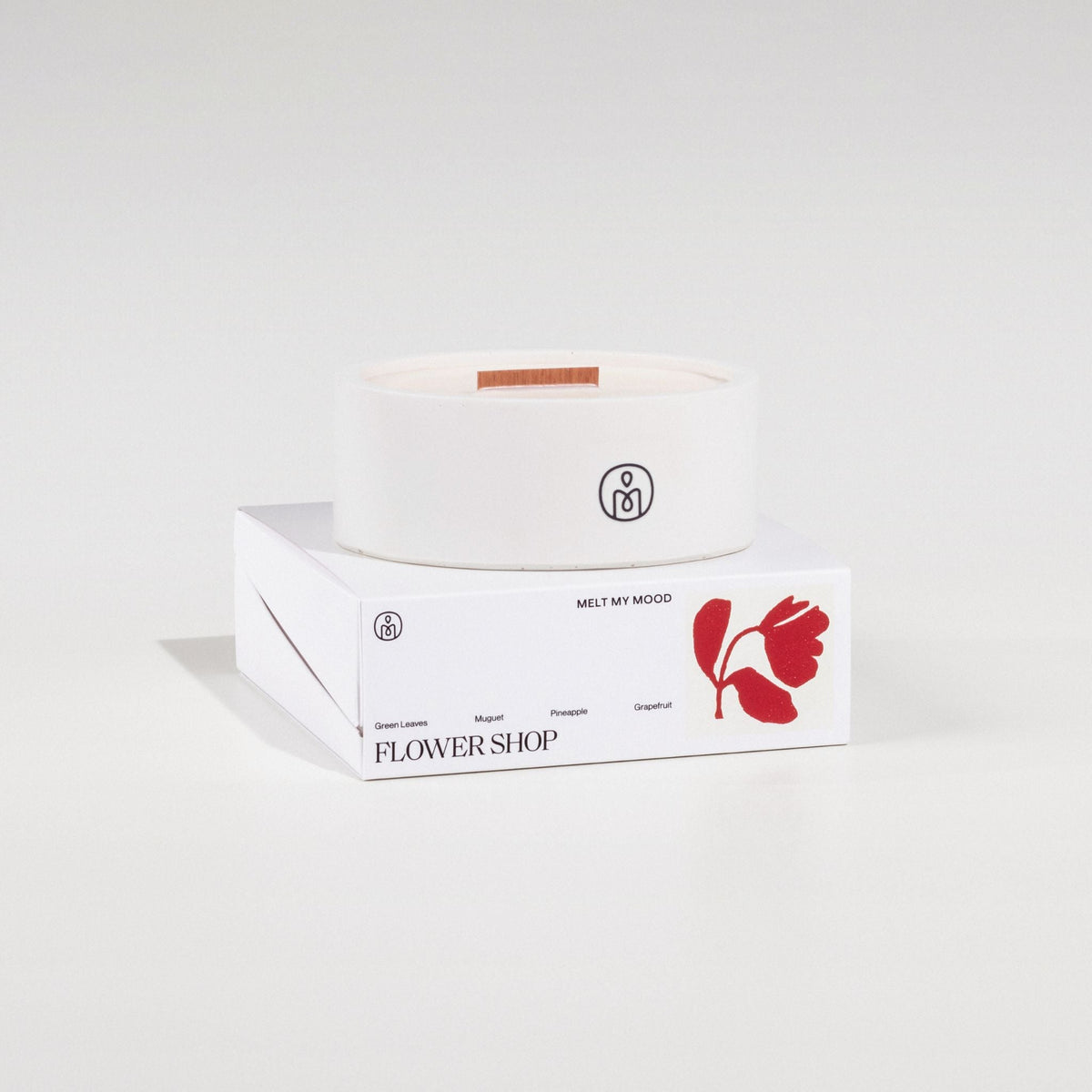 Egy kerek, törtfehér gyertya, benne fa kanóccal, egy fehér, téglalap alakú dobozon áll. A dobozon a „FLOWERSHOP” (a gyertya neve) és egy piros virág ikon látható; a felvétel tiszta fehér stúdióháttér előtt, lágy árnyékkal készült.