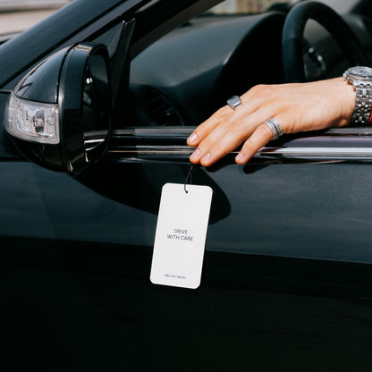 Egy „DRIVE WITH CARE” feliratú, fehér, téglalap alakú címke lóg az autó ajtókeretéről; a címkét egy fekete autó ablakpárkányán nyugvó kéz tartja.
