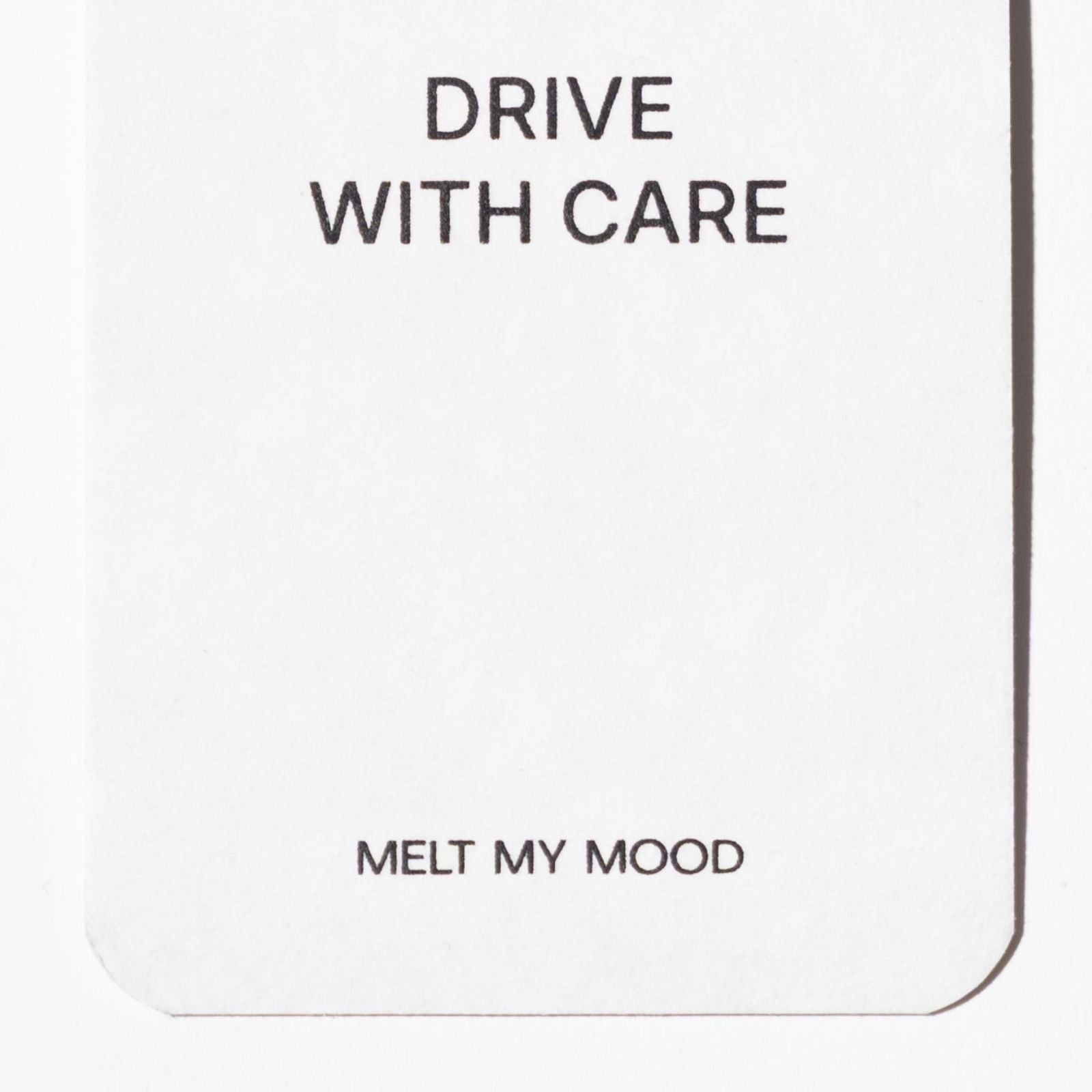 Lekerekített alsó sarkú, fehér, téglalap alakú autóillatosító; felső részén a „DRIVE WITH CARE”, alsó részéhez közel pedig a „MELT MY MOOD” felirat látható fekete betűkkel.
