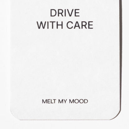 Lekerekített alsó sarkú, fehér, téglalap alakú autóillatosító; felső részén a „DRIVE WITH CARE”, alsó részéhez közel pedig a „MELT MY MOOD” felirat látható fekete betűkkel.
