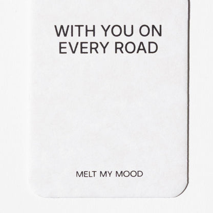 Lekerekített alsó sarkú, fehér, téglalap alakú autóillatosító; felső részén a „WITH YOU ON EVERY ROAD”, alsó részéhez közel pedig a „MELT MY MOOD” felirat látható fekete betűkkel.