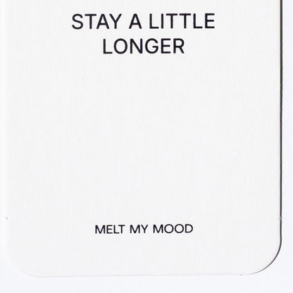 Közelkép egy lekerekített sarkú, fehér, téglalap alakú gardróbillatosítóról, amelynek felső részén a „STAY A LITTLE LONGER”, alsó részén pedig a „MELT MY MOOD” felirat látható fekete betűkkel.
